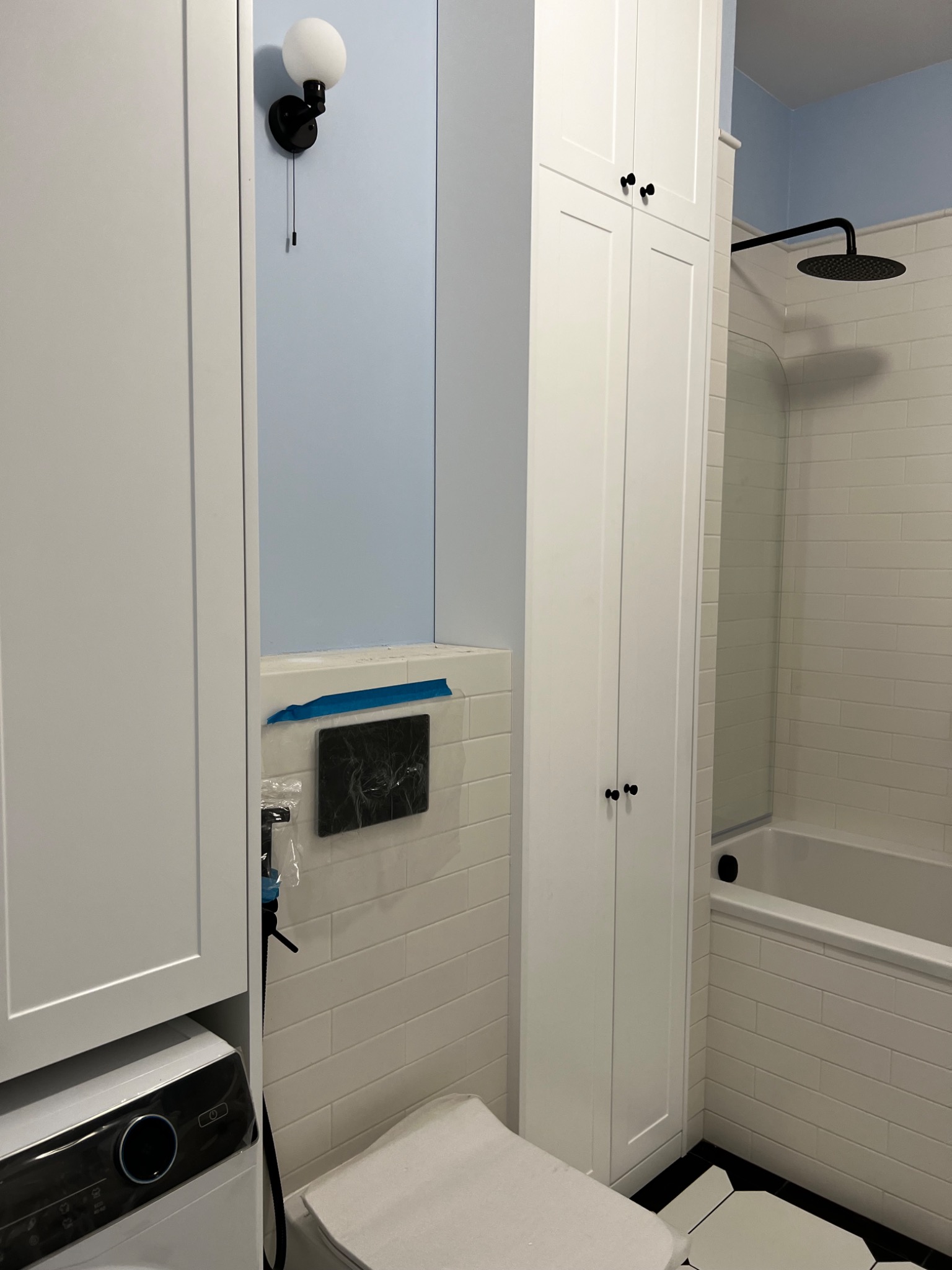 Nowoczesna łazienka z białą zabudową meblową, podtynkową spłuczką zabezpieczoną folią, kabiną prysznicową typu walk-in i czarno-białą podłogą.