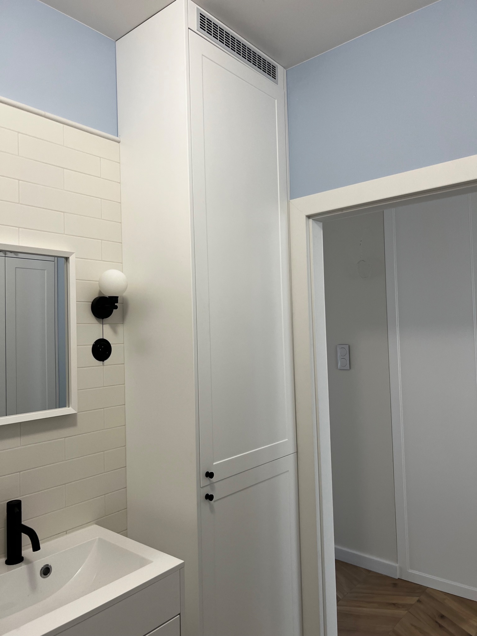 Nowoczesna łazienka z białą zabudową meblową na wymiar, umywalką z czarną armaturą i minimalistycznym oświetleniem ściennym.