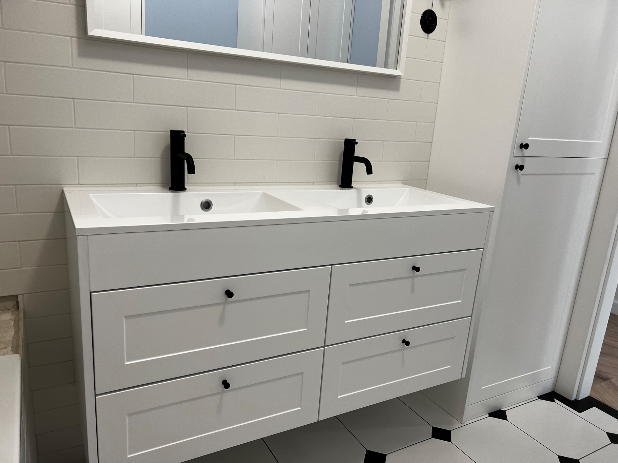 Biała szafka łazienkowa z dwiema umywalkami i czarnymi bateriami, uzupełniona białą szafką stojącą i czarno-białą podłogą w romby.