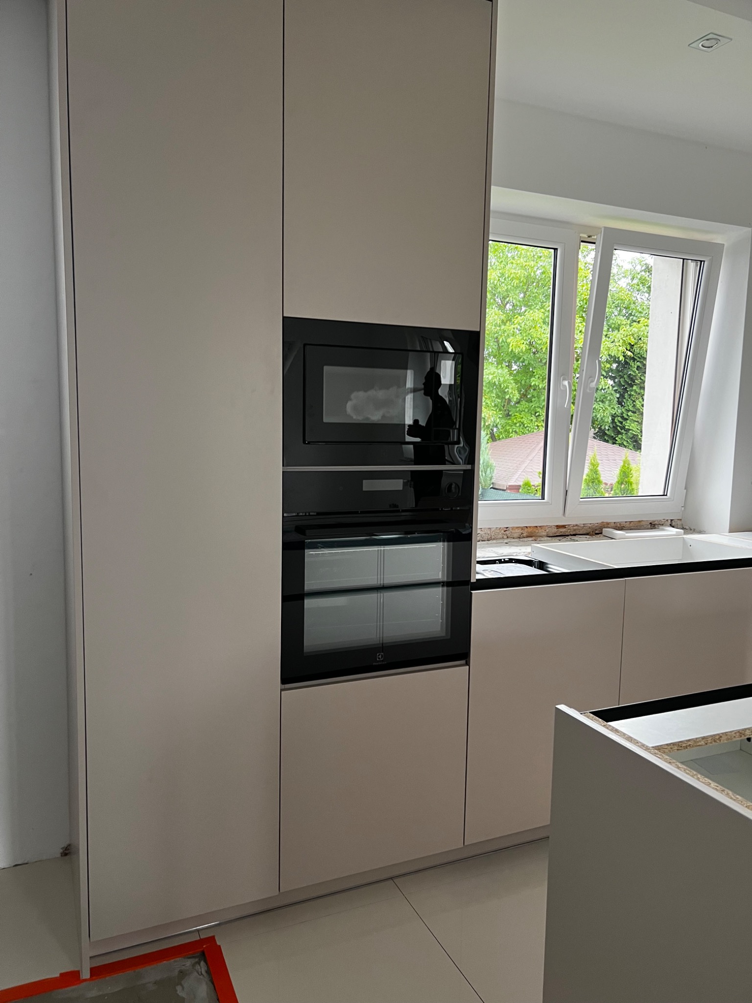 Beżowa zabudowa kuchenna z wbudowanym piekarnikiem i mikrofalówką, widok na okno z zielenią, fragment niedokończonego montażu blatu.