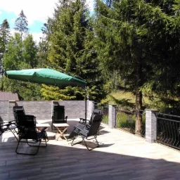 Taras z drewnopodobną podłogą, otoczony kamiennym murem i ozdobną metalową balustradą, wyposażony w czarne, rozkładane krzesła, mały drewniany stolik i duży zielony parasol, z widokiem na zalesione...