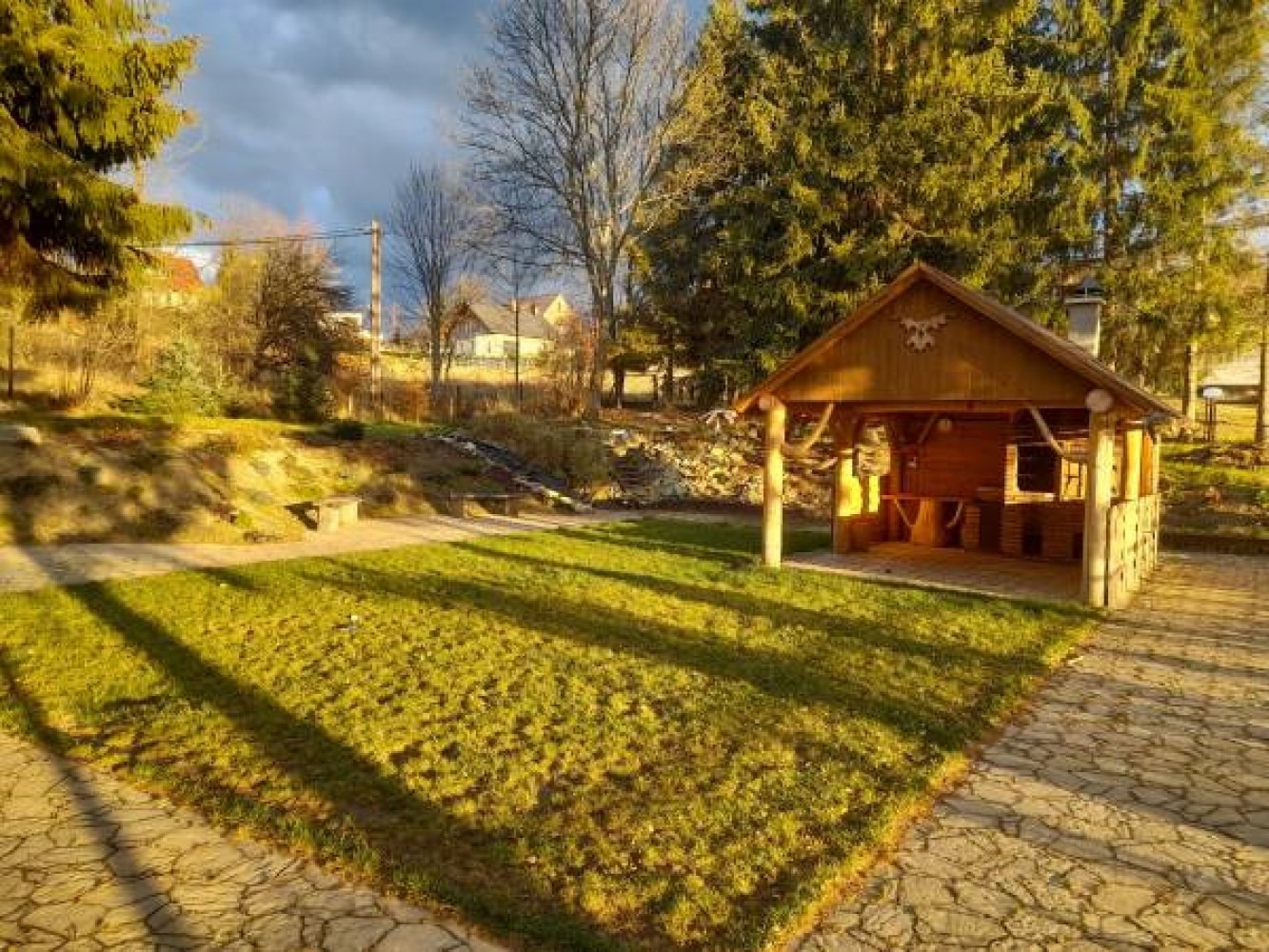 Drewniana altana grillowa z kominem w ogrodzie, oświetlona popołudniowym słońcem, z widokiem na pagórkowaty teren i domy w tle.