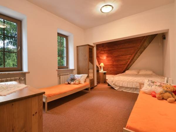 Pokój z trzema łóżkami: dwa pojedyncze z pomarańczowymi materacami, jedno podwójne pod drewnianą ścianą, jasne wnętrze z dwoma oknami i szafą, pluszowy miś na jednym z łóżek.
