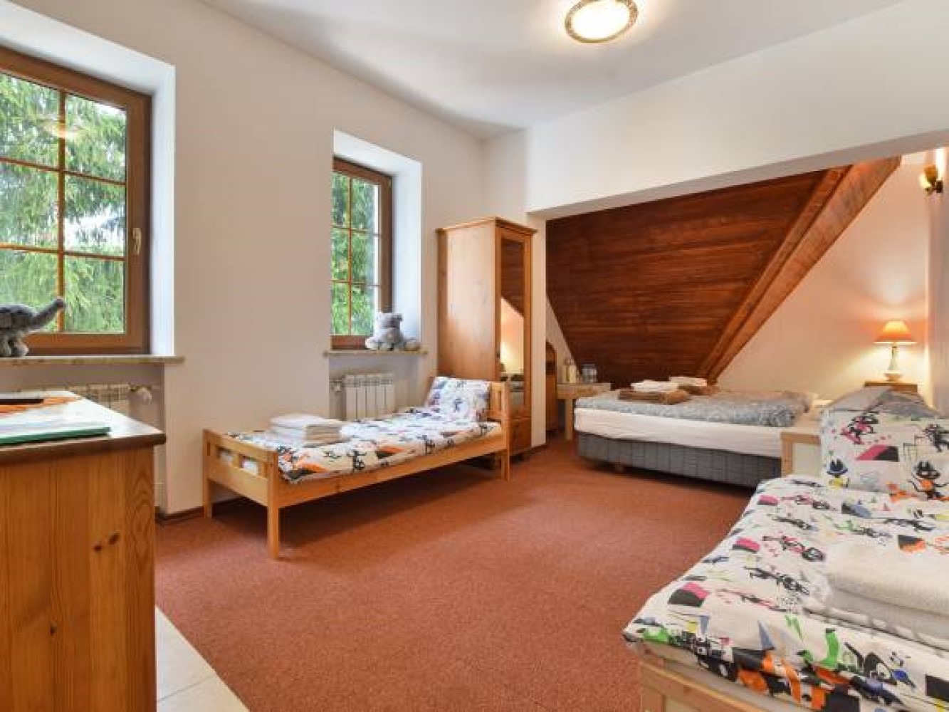 Pokój z trzema łóżkami, drewnianą szafą z lustrem, oknami z widokiem na drzewa, i skośnym drewnianym sufitem w tle. Na parapecie okiennym pluszowy słoń i miś.