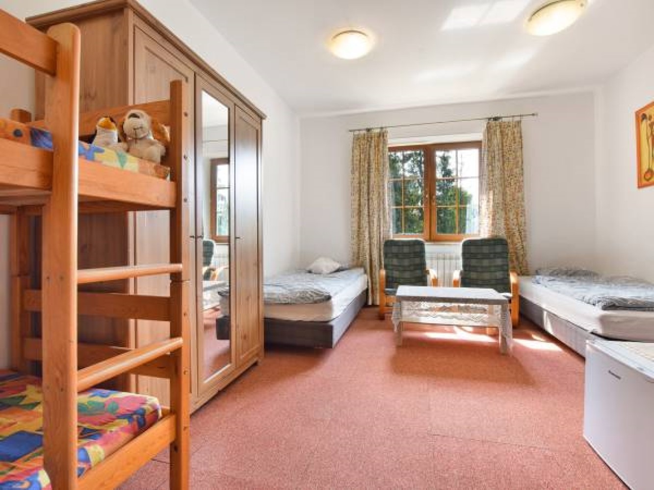 Pokój z łóżkiem piętrowym z pluszakami na górze, szafą z lustrem, dwoma pojedynczymi łóżkami, stolikiem i dwoma krzesłami przy oknie z zasłonami.