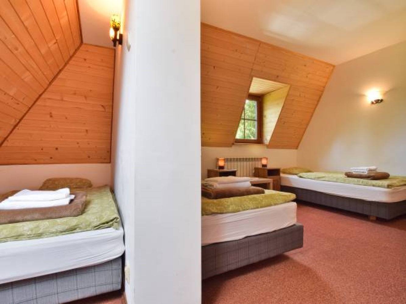 Pokój trzyosobowy na poddaszu z drewnianą boazerią, dwoma łóżkami pojedynczymi i małym oknem z widokiem na zieleń.