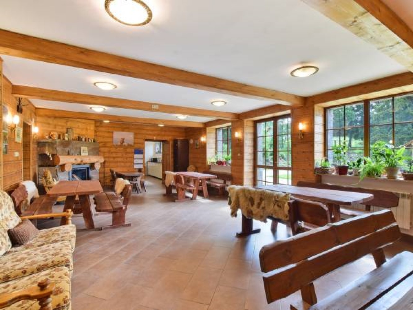 Duża jadalnia w stylu rustykalnym z drewnianymi ścianami, stołami z ławkami, kominkiem i widokiem na zieleń za oknami.