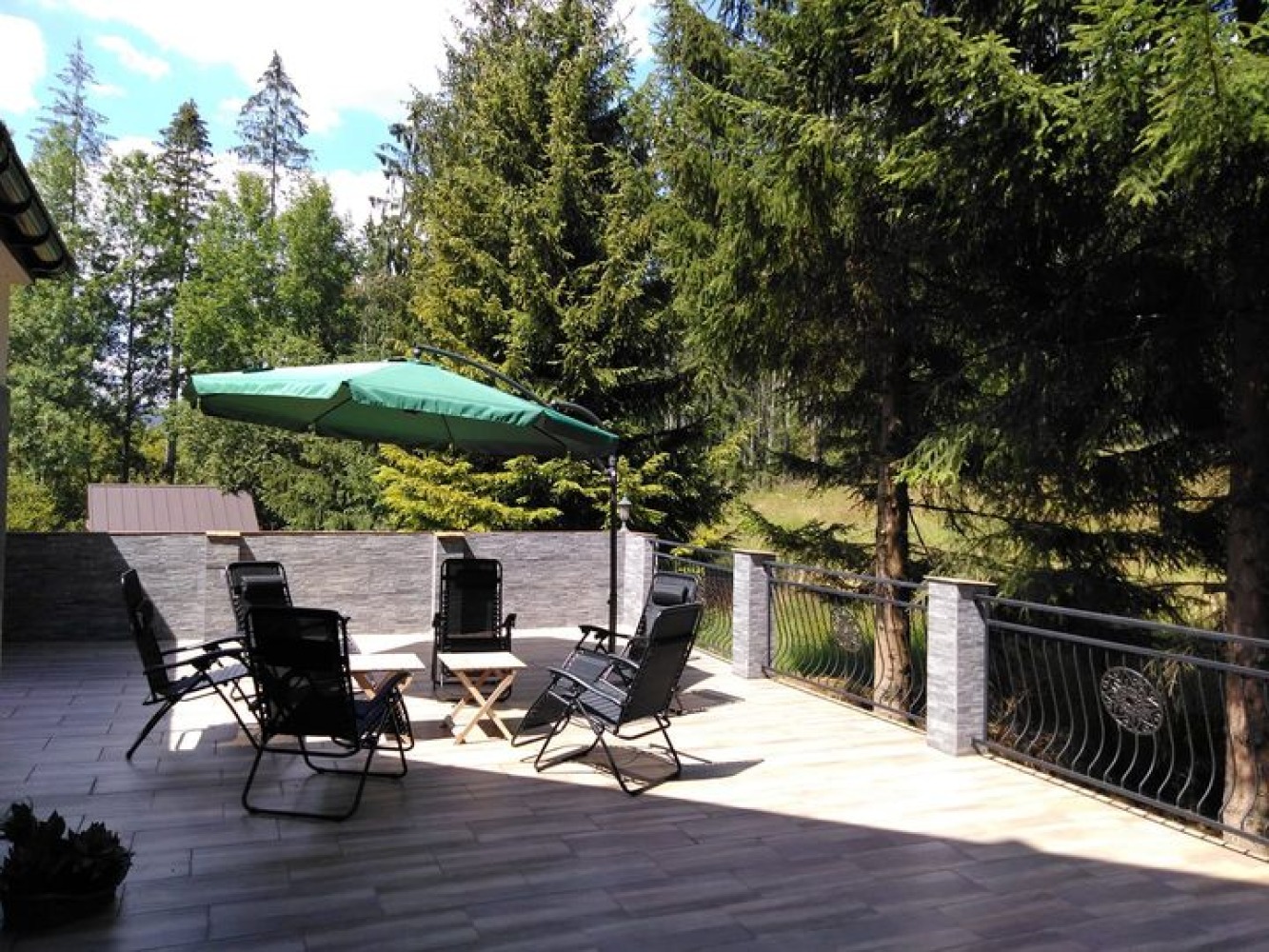 Taras z drewnopodobną podłogą, otoczony kamiennym murem i ozdobną metalową balustradą, wyposażony w czarne, rozkładane krzesła, mały drewniany stolik i duży zielony parasol, z widokiem na zalesione...