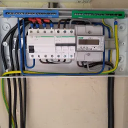 Otwarta skrzynka elektryczna z zainstalowanymi bezpiecznikami, licznikiem energii i przewodami w różnych kolorach, zamontowana na pomalowanej ścianie.