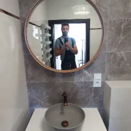 Szara umywalka nablatowa z brązową baterią w nowocześnie wykończonej łazience z marmurowymi płytkami; w lustrze odbicie instalatora robiącego zdjęcie telefonem.