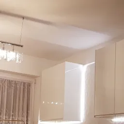 Nowoczesna kuchnia z białymi, połyskliwymi szafkami wiszącymi, oświetleniem LED pod szafkami i dekoracyjną lampą sufitową z kryształkami.