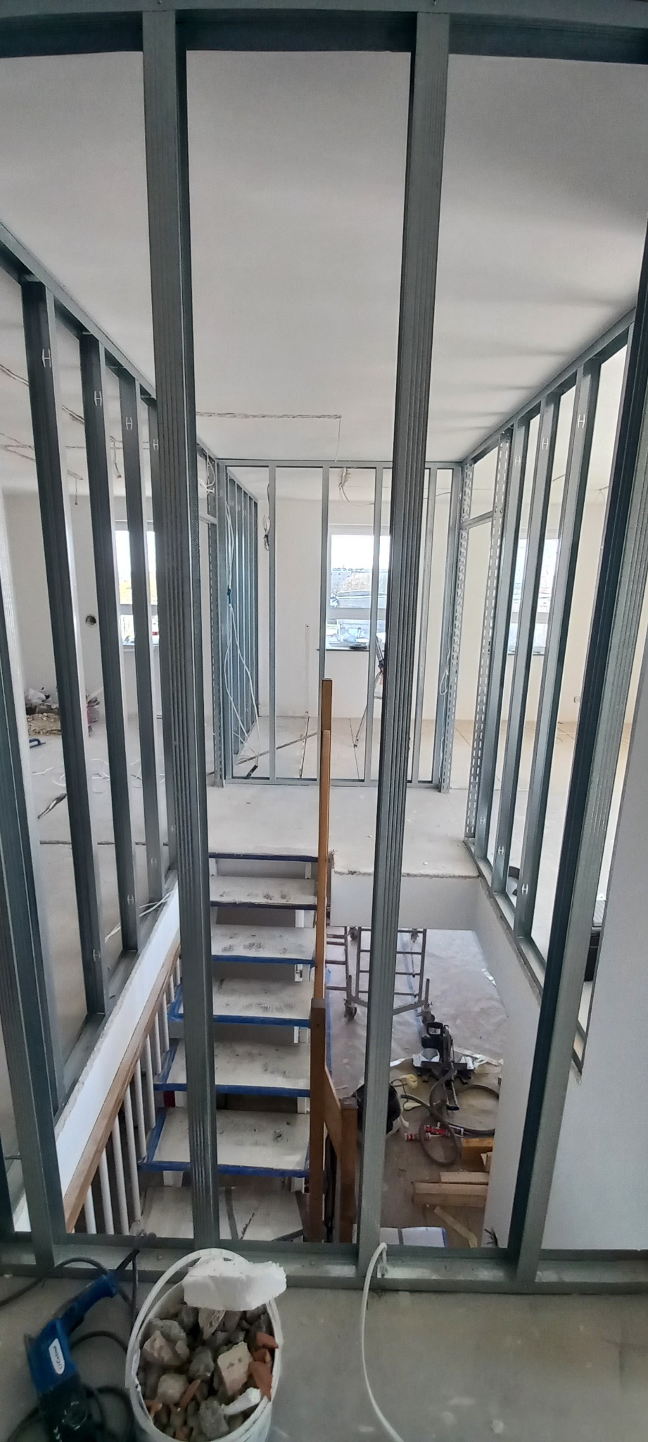 Wnętrze w trakcie remontu: widok z góry na metalową konstrukcję ścian działowych, schody z drewnianą barierką oraz narzędzia i gruz w wiadrze na podłodze.