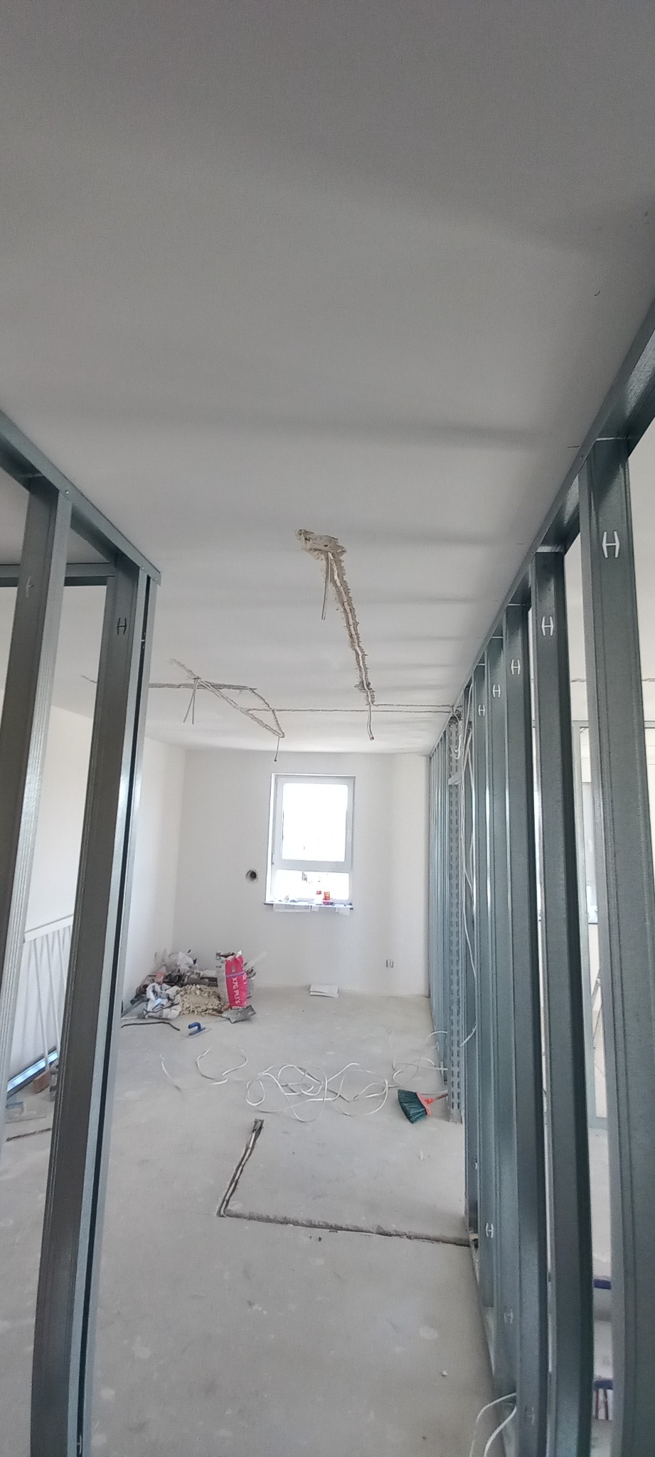 Wnętrze w trakcie remontu: widoczne metalowe profile ścian działowych, wystające przewody elektryczne z sufitu oraz kable na podłodze w surowym stanie.