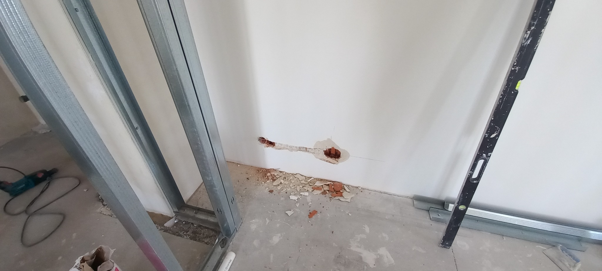 Przygotowanie ściany pod instalację: widoczne metalowe profile, wywiercony otwór w ścianie pod puszkę elektryczną z wystającymi przewodami, gruz na podłodze, poziomica oparta o ścianę.