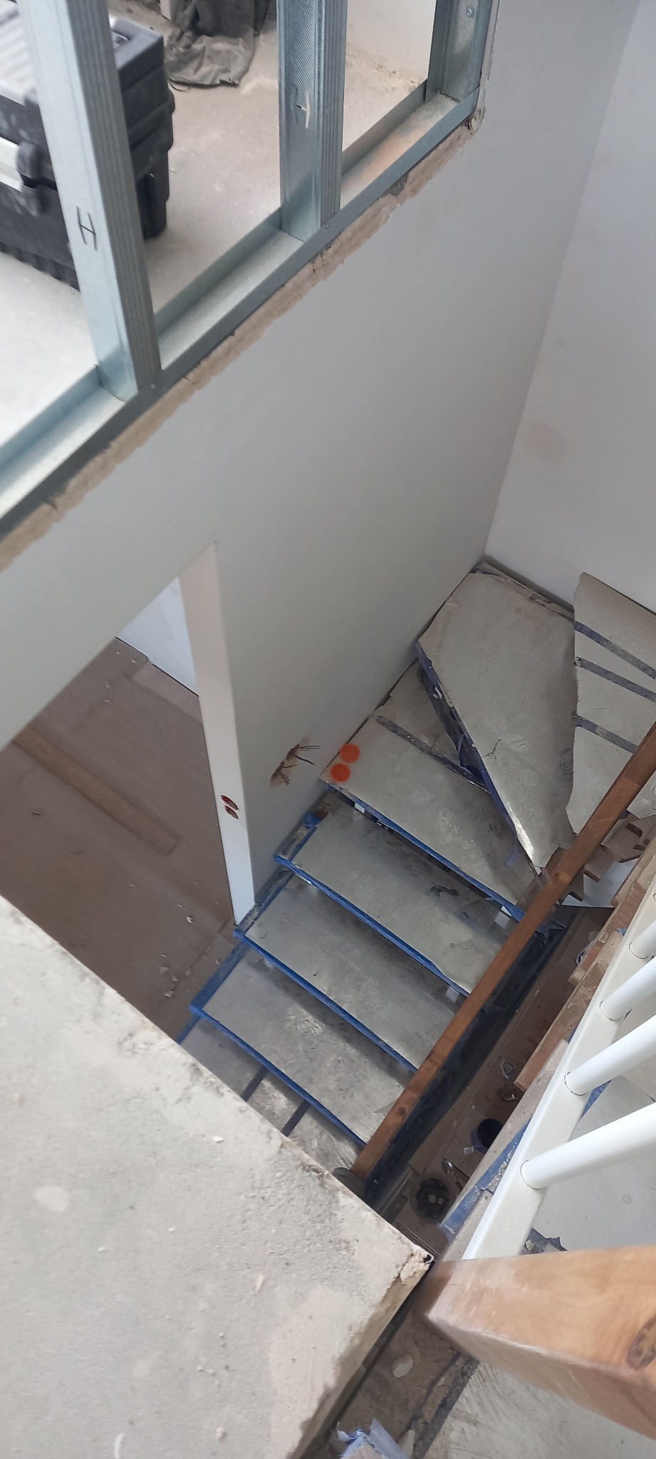 Widok z góry na surowe schody w trakcie wykańczania, z widocznymi przewodami elektrycznymi wystającymi ze ściany, metalowymi profilami przy oknie i częściowo ułożoną podłogą na dole.