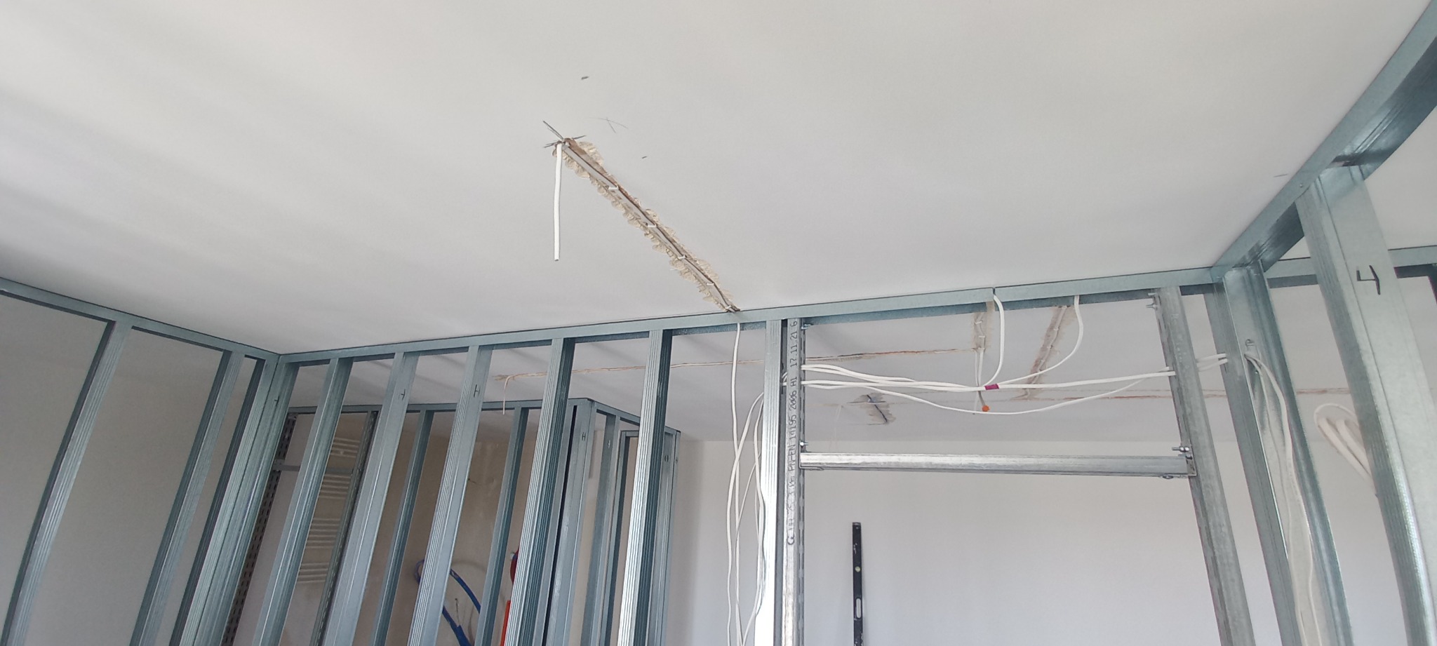 Wnętrze w trakcie remontu: metalowa konstrukcja ścian działowych, wystające przewody elektryczne z sufitu, widoczne ślady po kuciu w suficie.