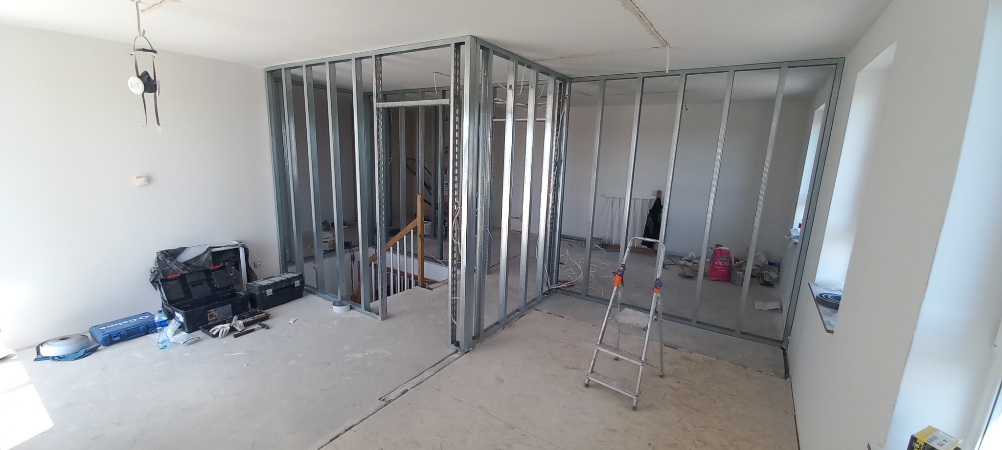 Wnętrze w trakcie budowy: metalowa konstrukcja szkieletowa ścian działowych, widoczne kable elektryczne, narzędzia i drabina, surowy beton na podłodze.