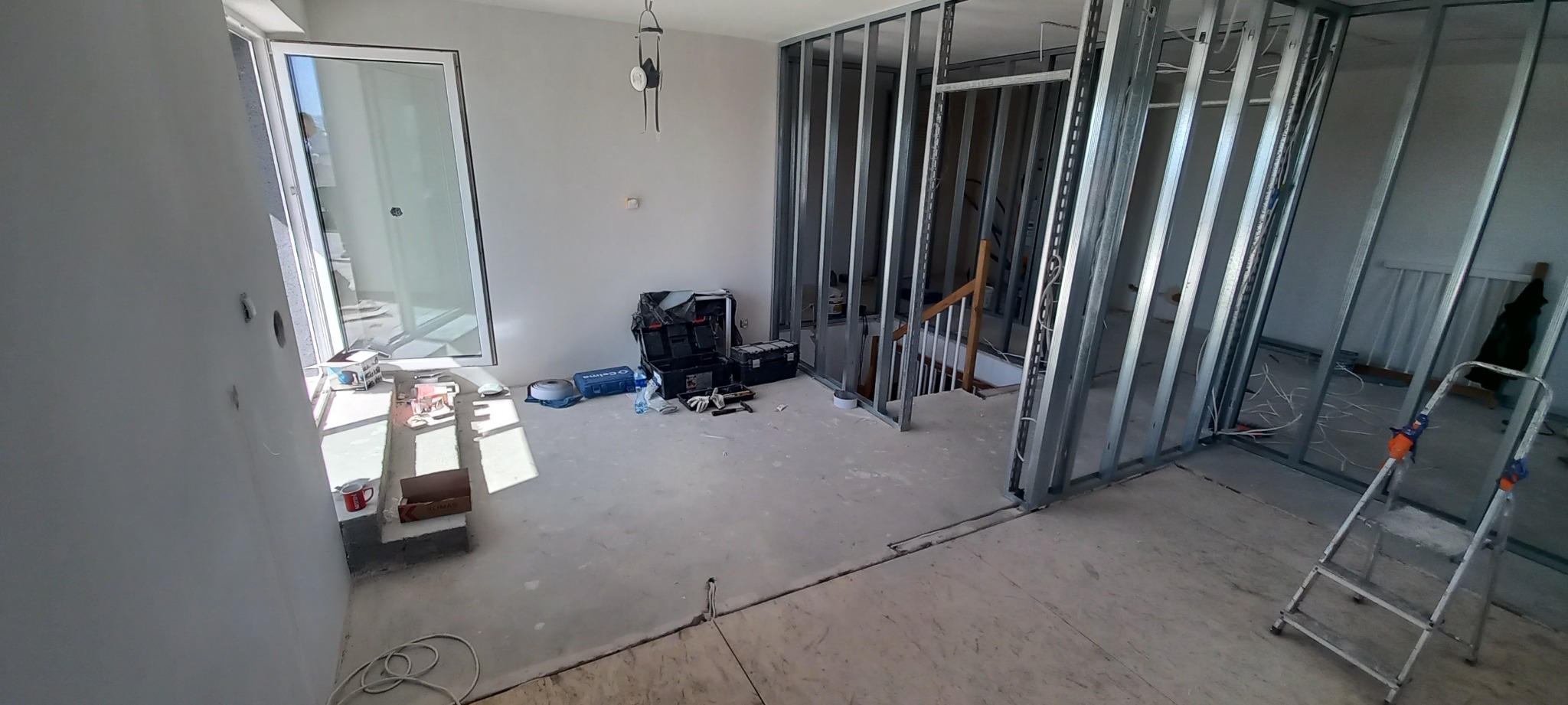 Wnętrze w trakcie instalacji ścian działowych z metalowych profili, widoczne narzędzia, skrzynki narzędziowe, przewody elektryczne wystające ze ściany, okno balkonowe z widokiem na zewnątrz, schody...