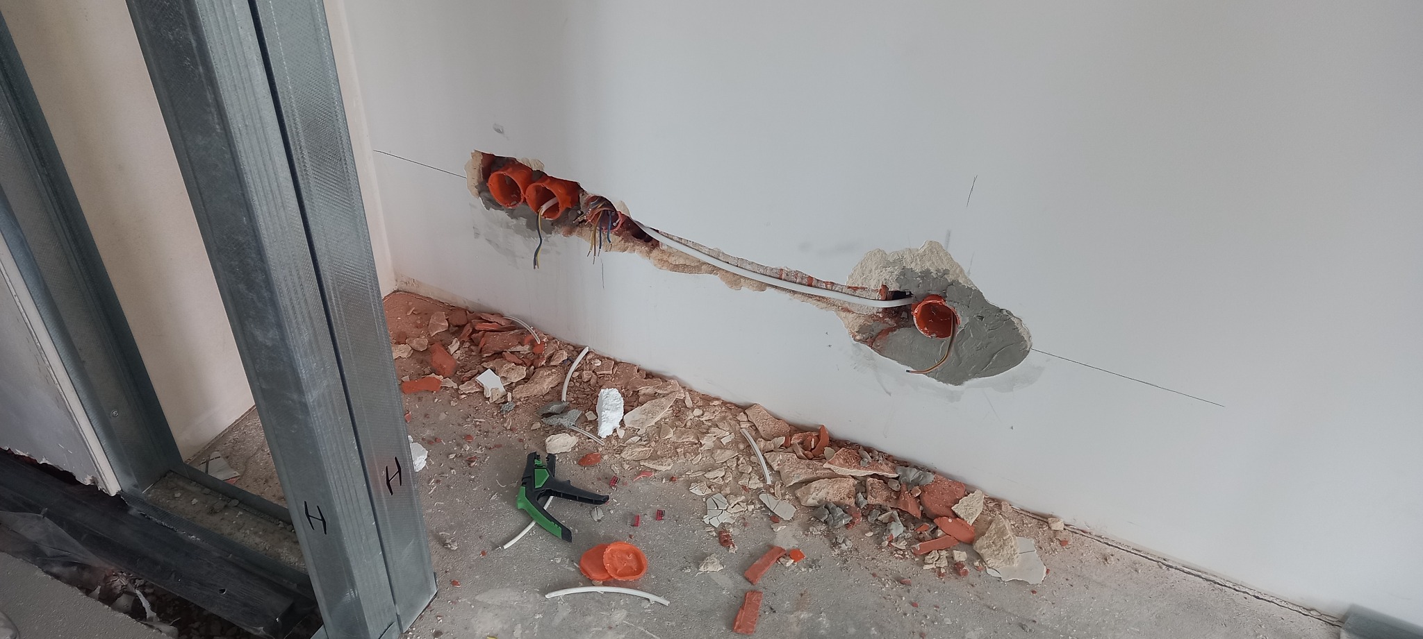 Wykute otwory w ścianie z przewodami elektrycznymi w pomarańczowych peszlach, widoczne narzędzia do zdejmowania izolacji i fragmenty gruzu na podłodze, metalowa konstrukcja ścianki działowej w tle.