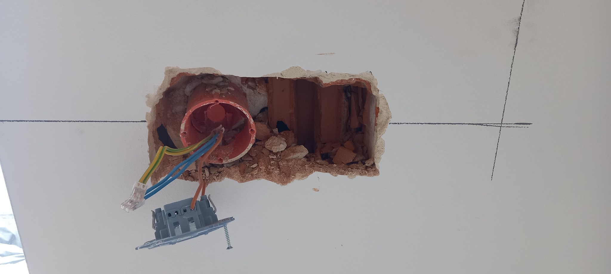 Otwór w ścianie z widoczną pomarańczową puszką instalacyjną, wystającymi przewodami i zamontowanym gniazdkiem w trakcie montażu, zarysowane linie pomocnicze na ścianie.