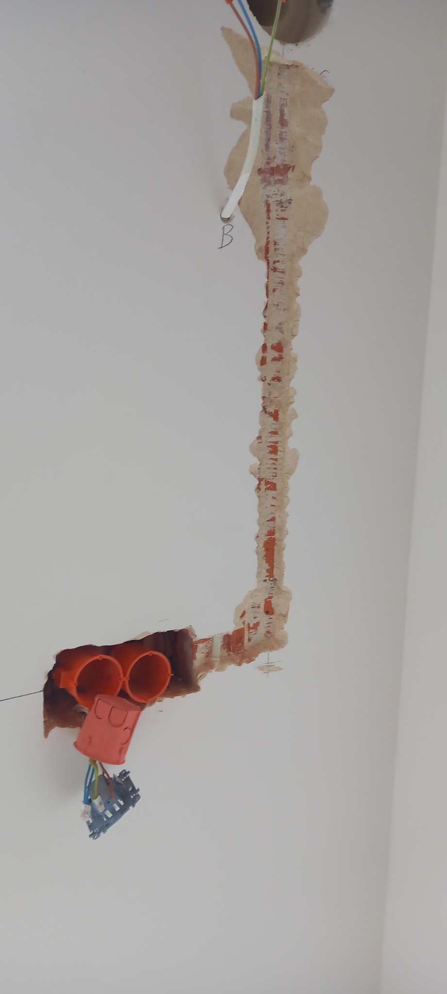 Wykuty w ścianie pionowy rowek pod instalację elektryczną z wystającymi przewodami, widoczne pomarańczowe rury instalacyjne w ścianie, puszka elektryczna z przewodami w dolnej części ściany, ślady...