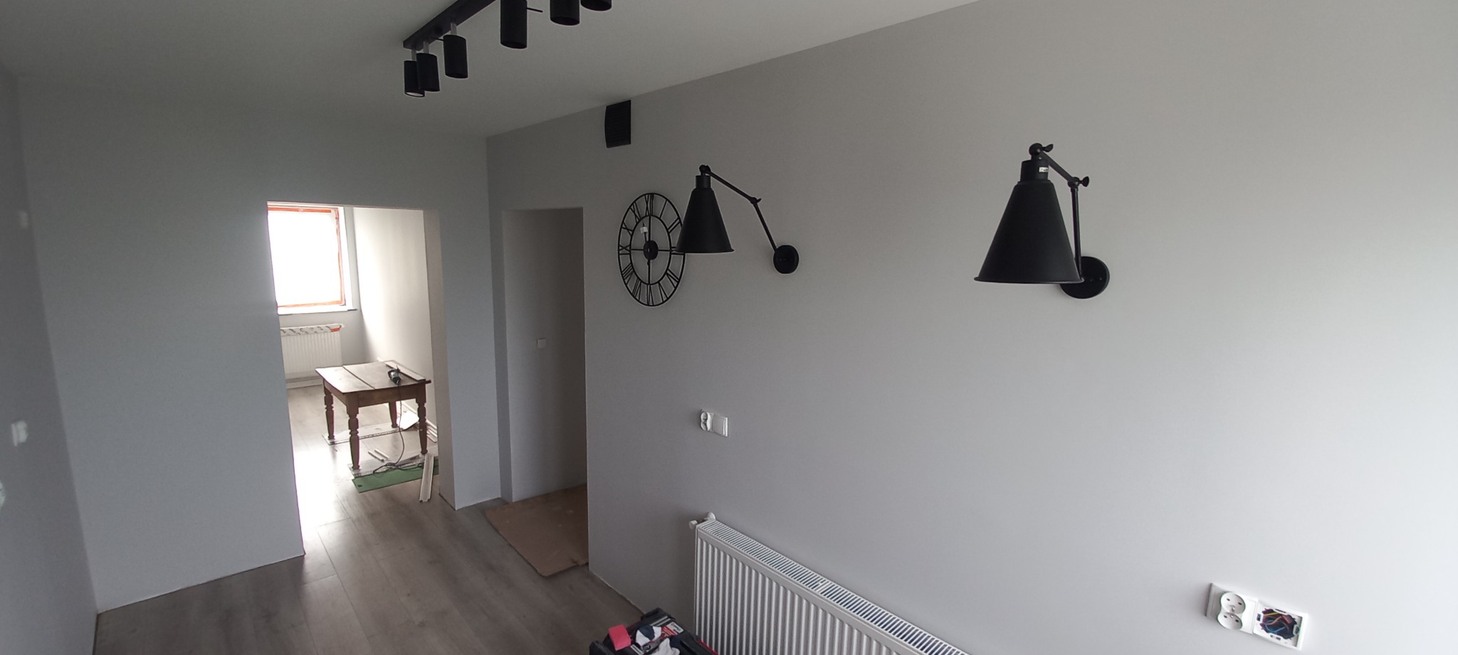 Szare wnętrze z czarnymi lampami ściennymi w stylu industrialnym, widoczne gniazdka elektryczne w trakcie montażu i narzędzia w tle.