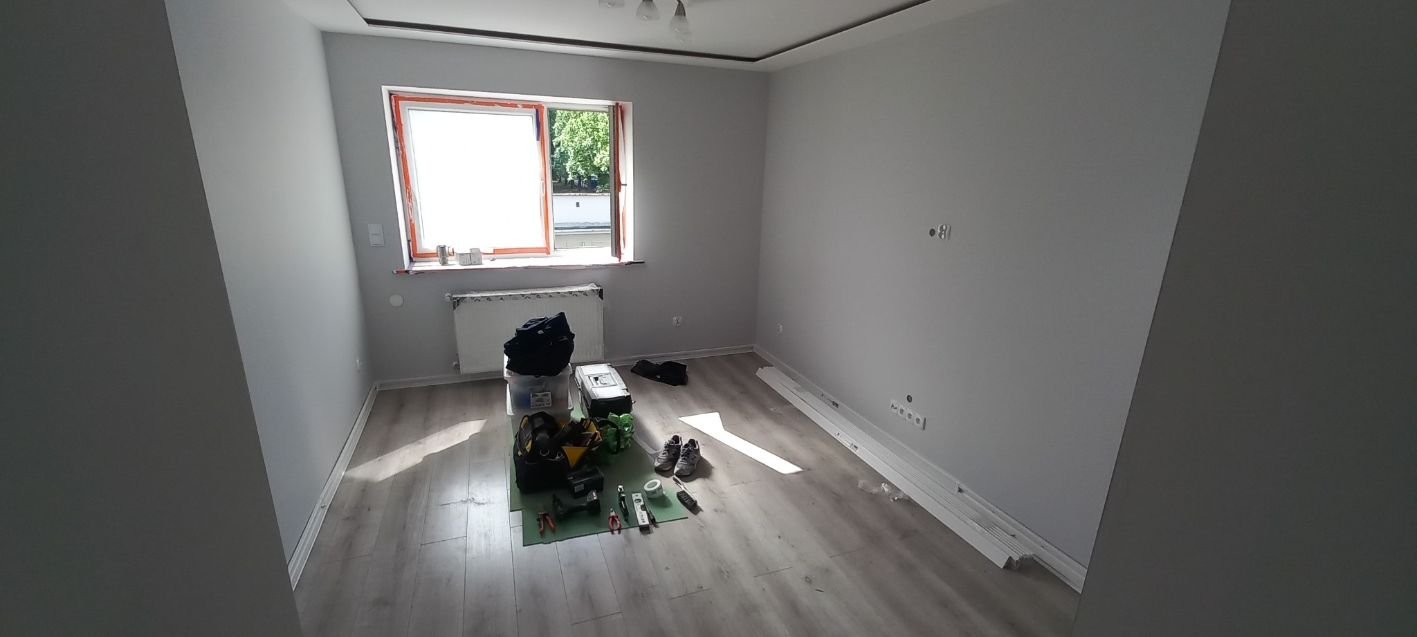 Wnętrze szarego pokoju z narzędziami elektrycznymi na podłodze, widoczne gniazdka i puszki instalacyjne na ścianie, listwy przypodłogowe przygotowane do montażu.