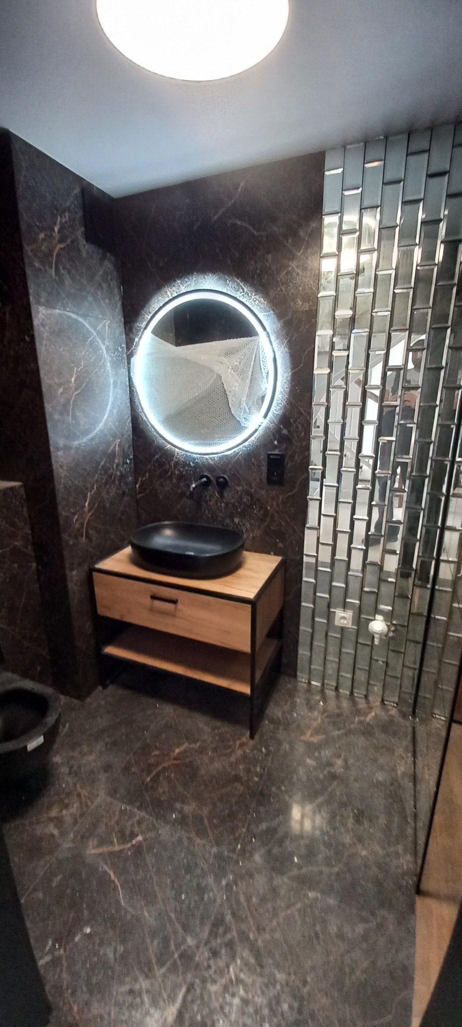 Nowoczesna łazienka z umywalką nablatową w kolorze czarnym, okrągłym lustrem z podświetleniem LED i ścianą wyłożoną mozaiką z małych lusterek.