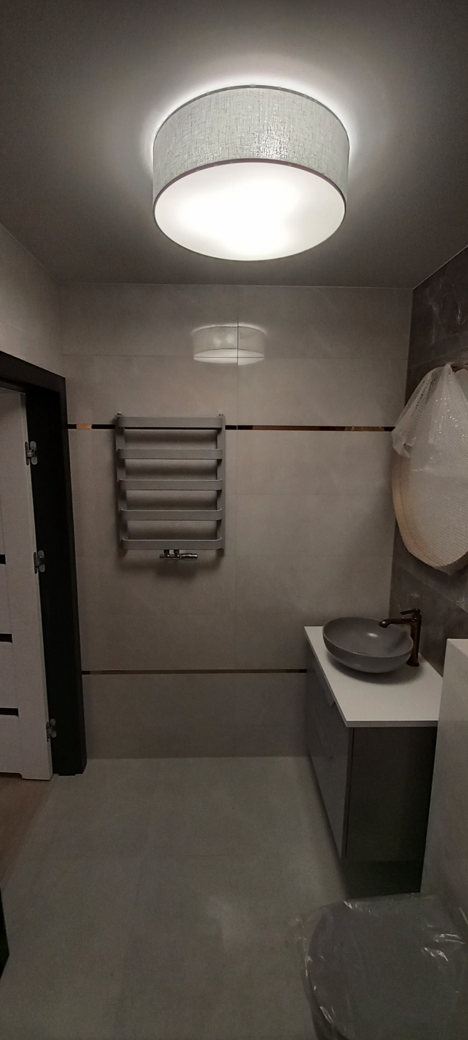 Nowoczesna łazienka z okrągłą lampą sufitową, umywalką nablatową w odcieniach szarości i grzejnikiem drabinkowym na ścianie z płytek w neutralnej kolorystyce.