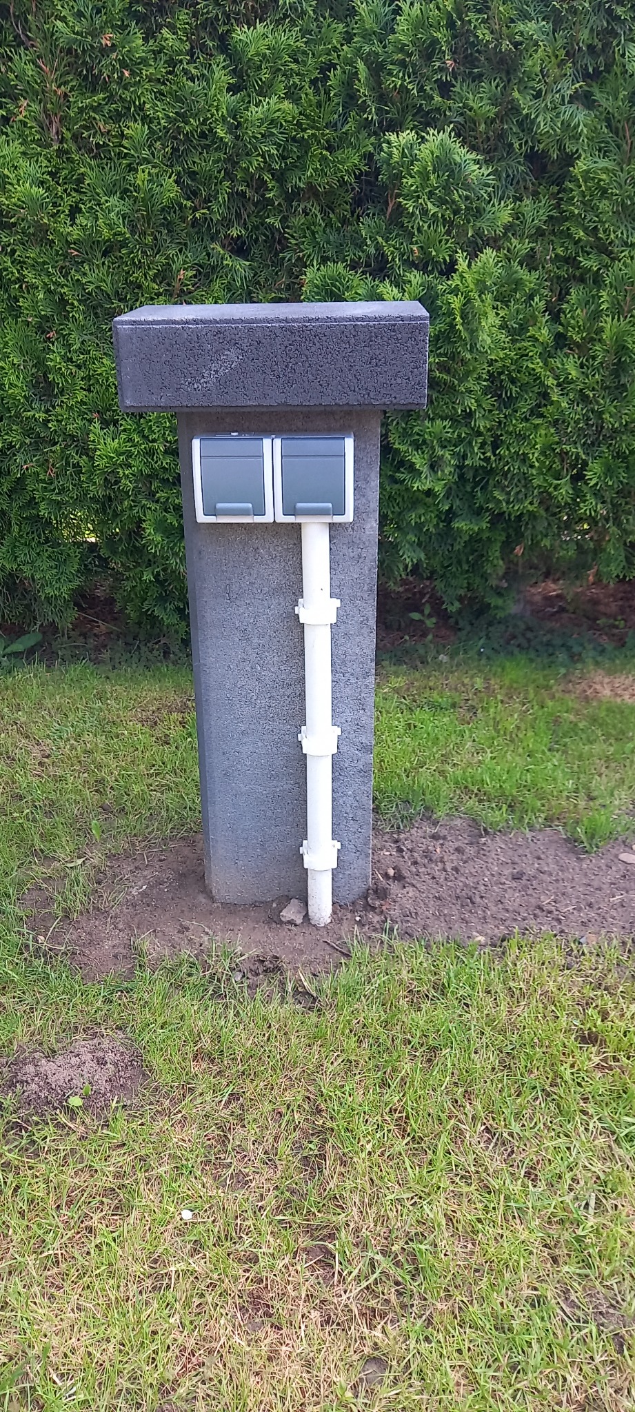 Zewnętrzna skrzynka elektryczna z dwoma gniazdami, umieszczona na betonowym słupku w ogrodzie, z widoczną białą rurą osłonową na przewody.