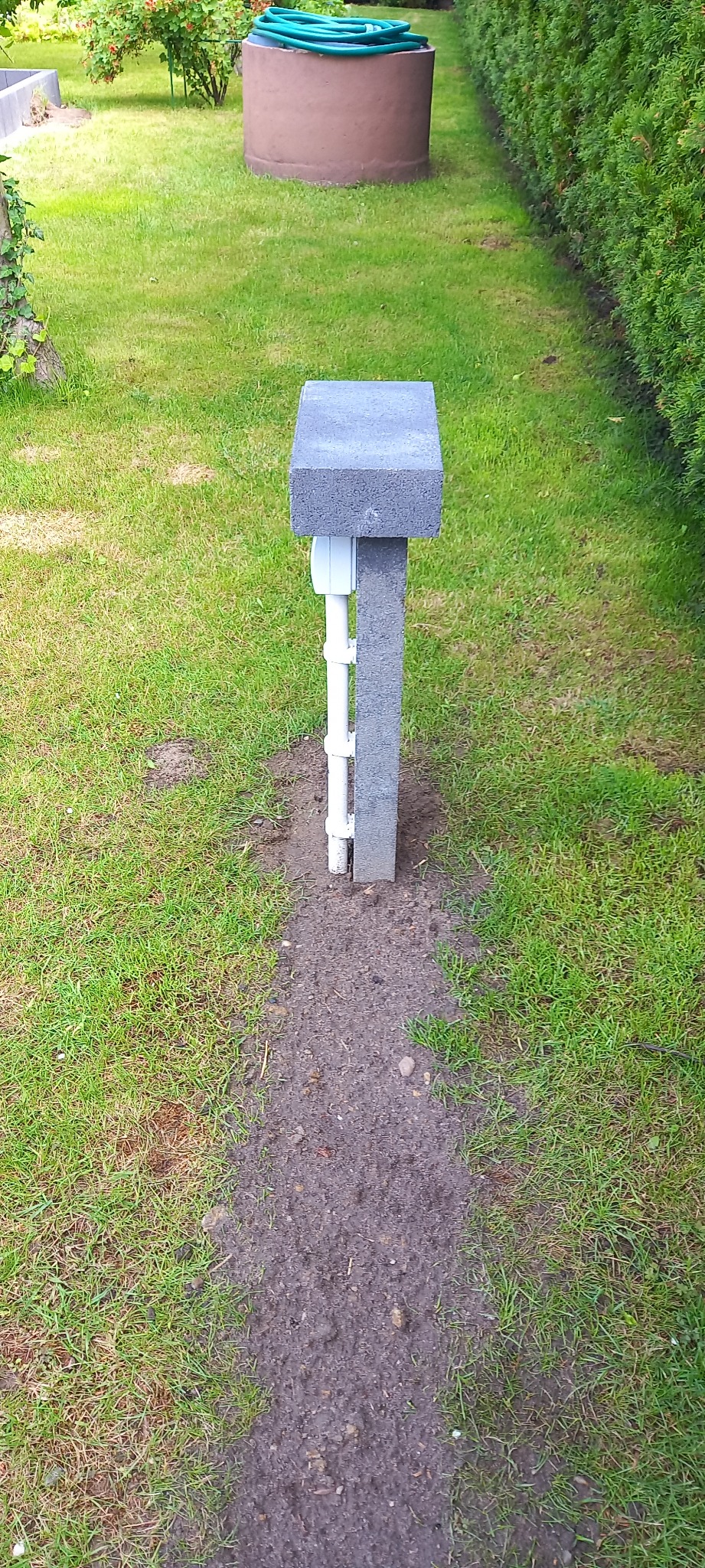 Zewnętrzna skrzynka elektryczna z betonową obudową i białymi rurami PCV na trawniku, widoczne zielone trawniki i wysokie tuje w tle.