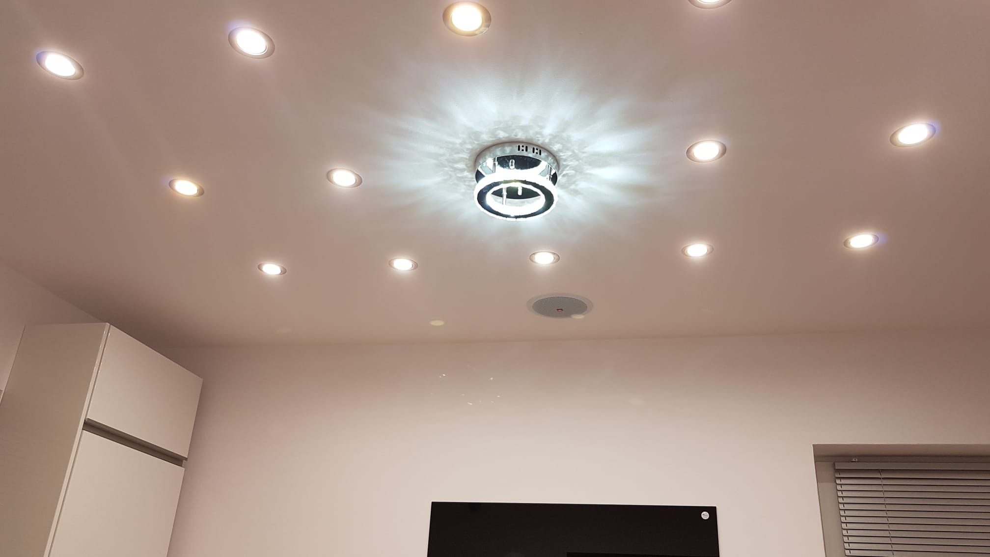 Sufit z równomiernie rozmieszczonymi punktowymi lampami LED i centralnym, okrągłym żyrandolem z chromowanymi pierścieniami, oświetlający białe ściany i fragment zabudowy kuchennej.