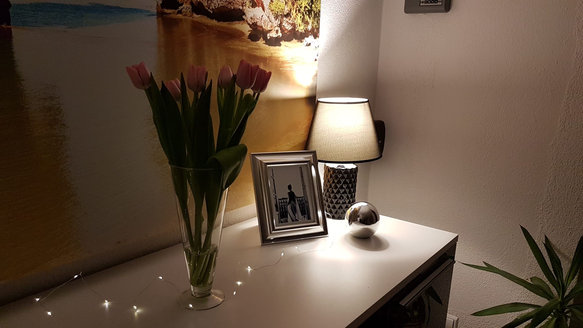 Ustawienie na białej komodzie: bukiet różowych tulipanów w szklanym wazonie, srebrna ramka ze zdjęciem, lampka nocna z abażurem, dekoracyjna kula i delikatne światełka, a w tle obraz z motywem plaży.