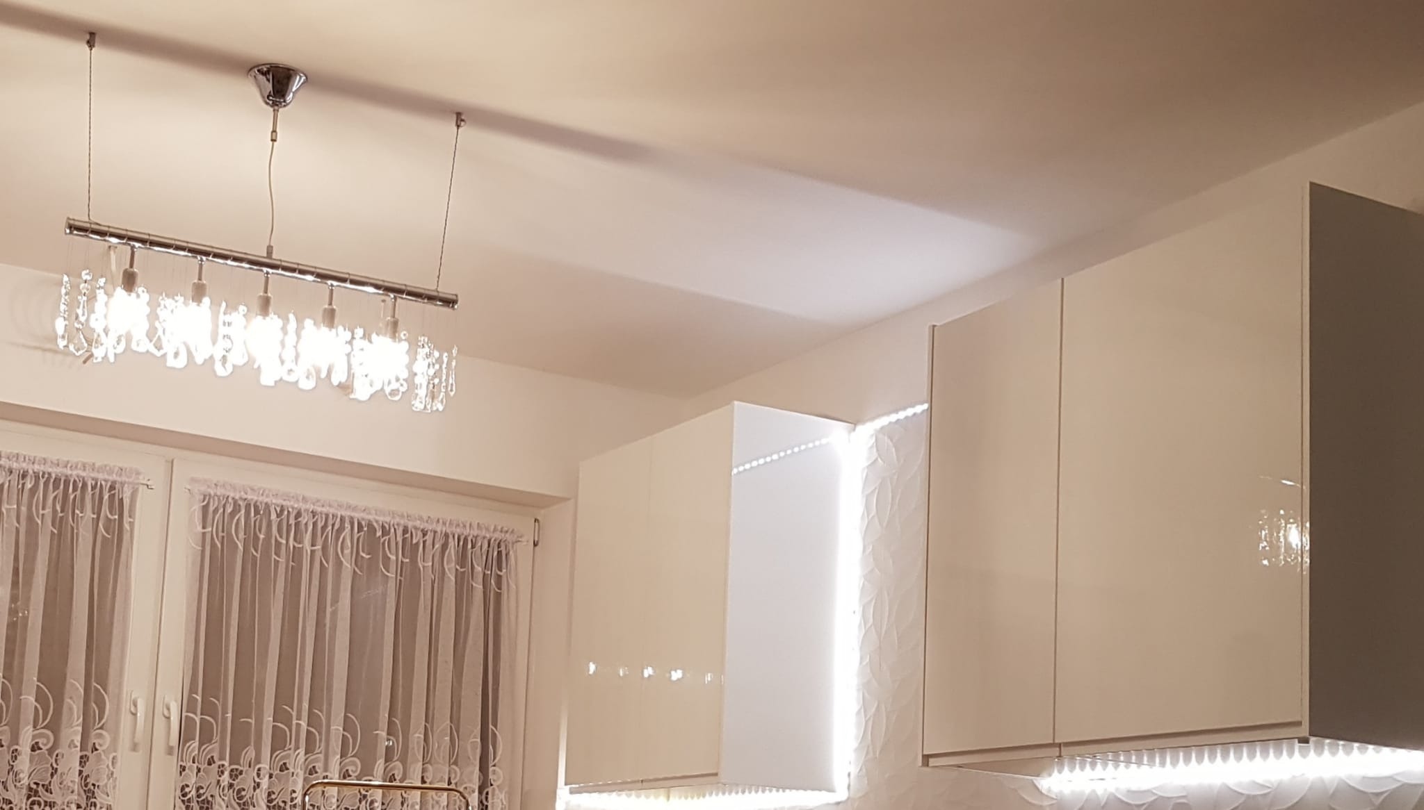 Nowoczesna kuchnia z białymi, połyskliwymi szafkami wiszącymi, oświetleniem LED pod szafkami i dekoracyjną lampą sufitową z kryształkami.