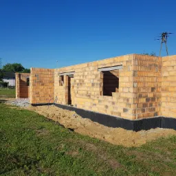 Wznoszone ściany parterowego domu z pustaków ceramicznych, widoczne otwory okienne i drzwiowe, zabezpieczone fundamenty czarną folią, w tle błękitne niebo i słup wysokiego napięcia.