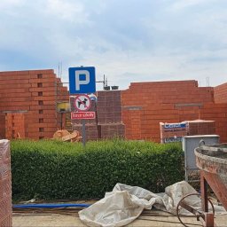 Volodymyr Kuvak - Wznoszenie murów z czerwonej cegły na placu budowy, widoczne znaki drogowe i materiały budowlane. Ujęcie pokazuje postęp prac murarskich w słoneczny dzień.