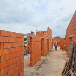 Volodymyr Kuvak - Wznoszenie ścian domu z czerwonej cegły, widok na budowę z perspektywy murarza, palety cegieł w tle. Robotnik w oddali przenosi materiały budowlane.