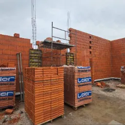 Wznoszenie ścian z czerwonej cegły Leier na placu budowy. Widoczne rusztowania, zbrojenie, palety z cegłami i fragment żurawia. Prace murarskie w toku.