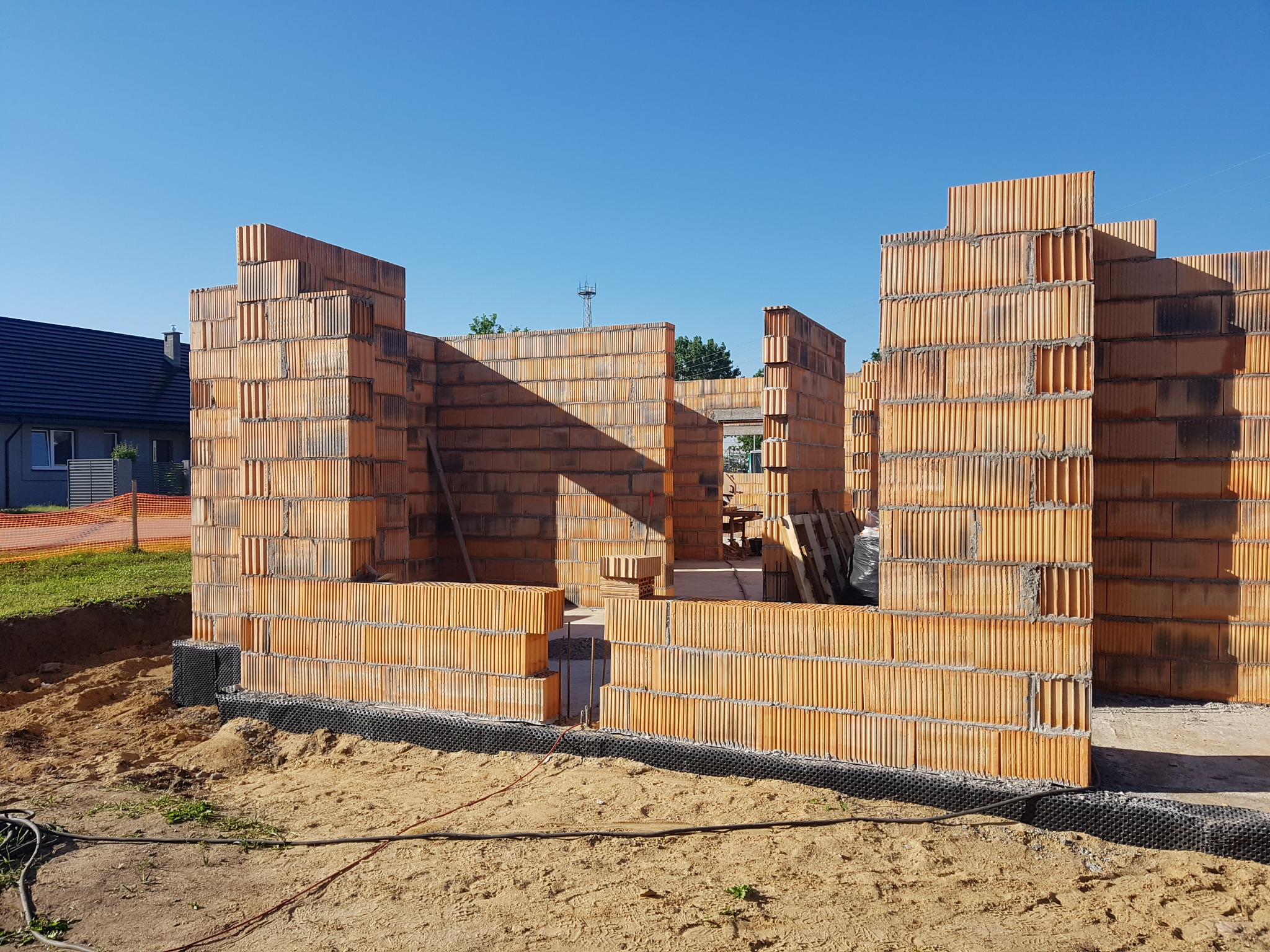 Wznoszone ściany domu z pustaków ceramicznych, widoczna czarna izolacja fundamentów, na zewnątrz piasek i trawa, w tle dom z ciemnym dachem i błękitne niebo.