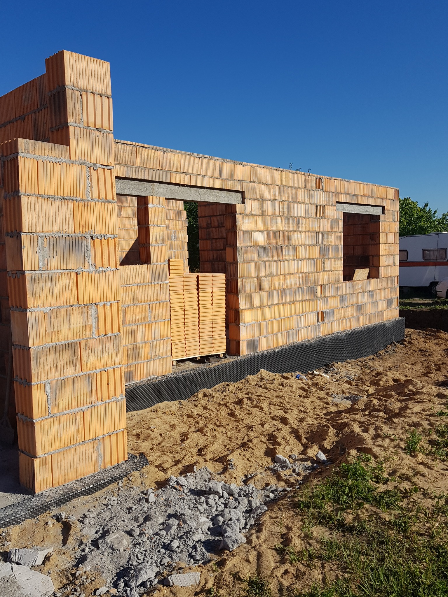 Wznoszona ściana domu z pustaków ceramicznych z widocznymi otworami okiennymi, zabezpieczona czarną folią hydroizolacyjną u podstawy, z paletą pustaków wewnątrz budynku i przyczepą kempingową w tle.