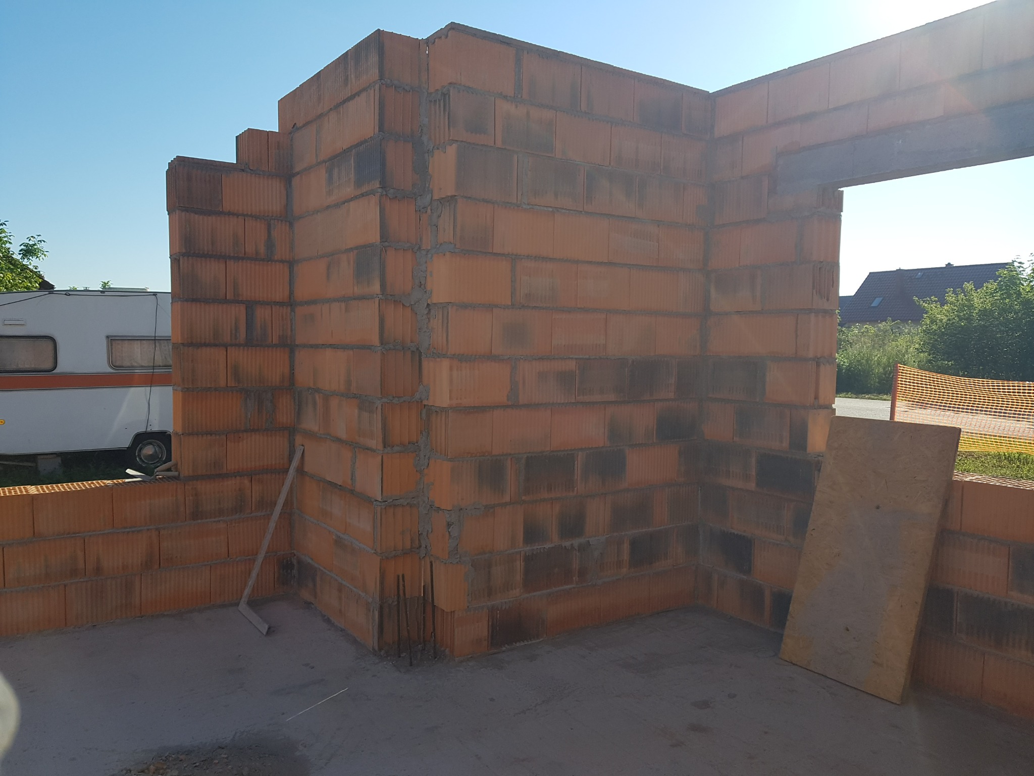 Wznoszenie ścian z czerwonej cegły ceramicznej, widoczny fragment muru z otworem okiennym, w tle przyczepa kempingowa i zieleń.