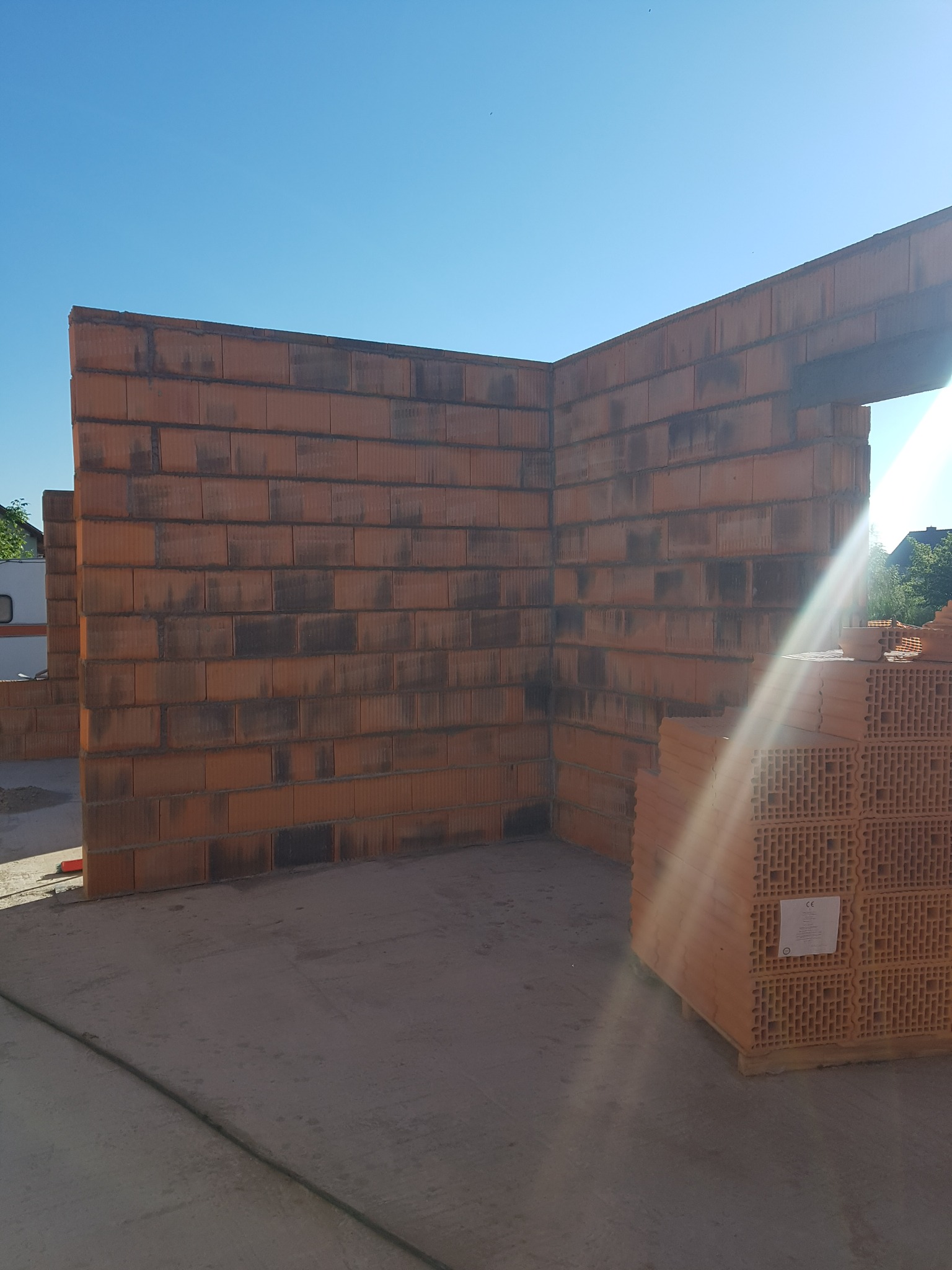 Wznoszone ściany z czerwonej ceramiki poryzowanej, widoczne palety z pustakami ceramicznymi, jasne światło słoneczne.