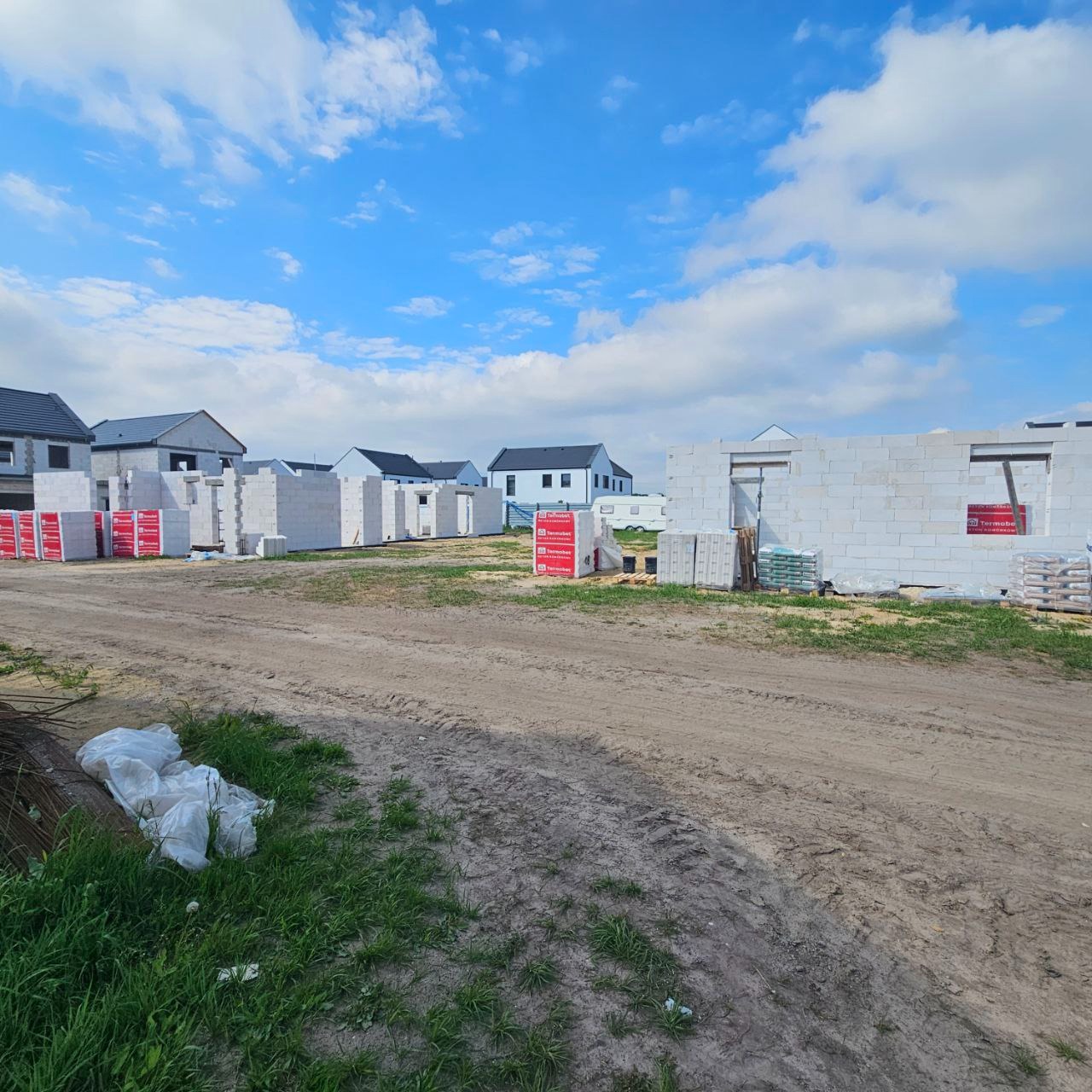 Budowa domów w trakcie realizacji: wzniesione ściany z bloczków, zapakowane materiały budowlane, domy w tle, słoneczny dzień, błękitne niebo z chmurami.