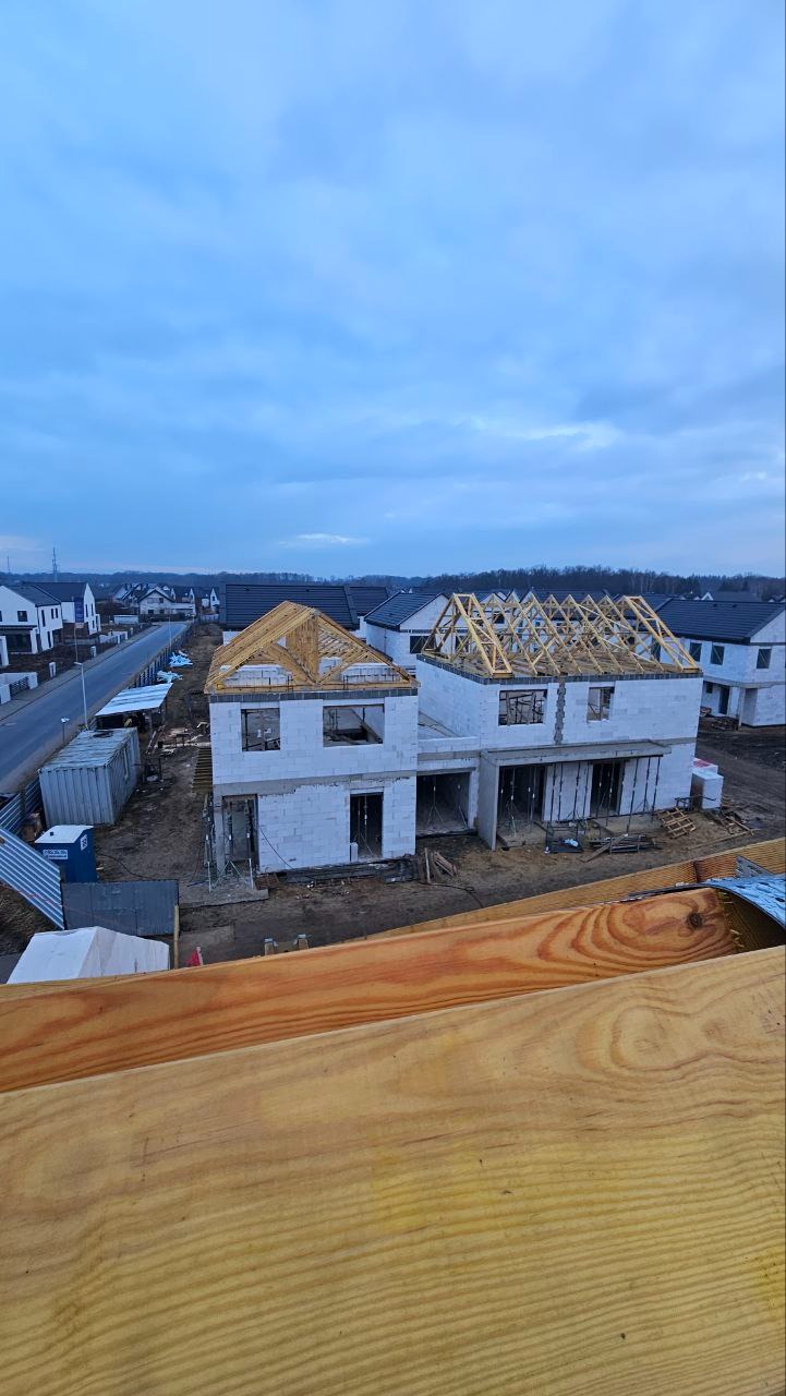 Budowa bliźniaka z białego betonu komórkowego, widok z góry na konstrukcję dachu, stan surowy otwarty, w tle zabudowa jednorodzinna i pochmurne niebo.