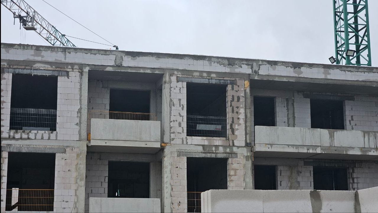 Wznoszenie ścian z betonu komórkowego, widoczne otwory okienne i balkonowe. Częściowo ukończona konstrukcja budynku wielorodzinnego z widocznymi żurawiami w tle.
