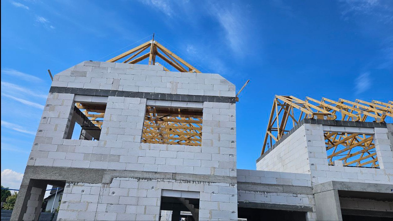 Surowy stan domu z betonu komórkowego, widoczne drewniane elementy konstrukcji dachu na tle błękitnego nieba. Ściany z betonu komórkowego z widocznymi otworami okiennymi.