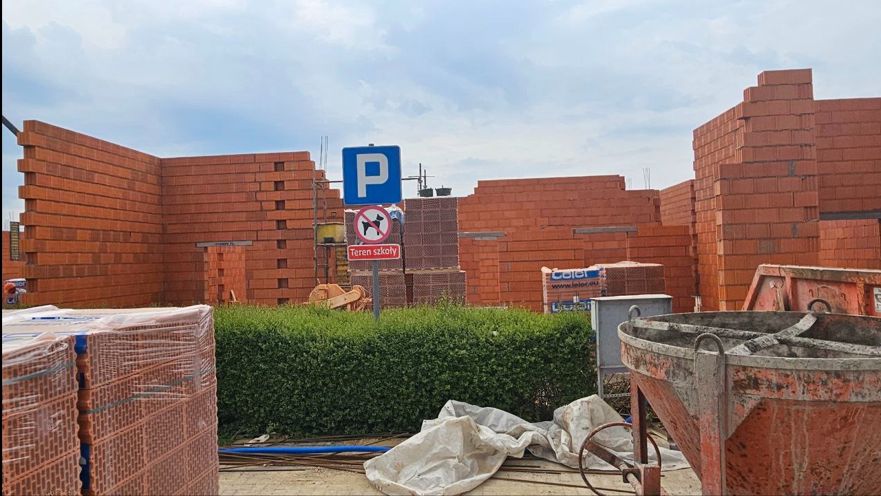 Wznoszenie murów z czerwonej cegły na placu budowy, widoczne znaki drogowe i materiały budowlane. Ujęcie pokazuje postęp prac murarskich w słoneczny dzień.