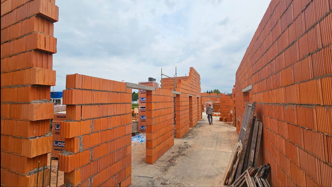 Wznoszenie ścian domu z czerwonej cegły, widok na budowę z perspektywy murarza, palety cegieł w tle. Robotnik w oddali przenosi materiały budowlane.