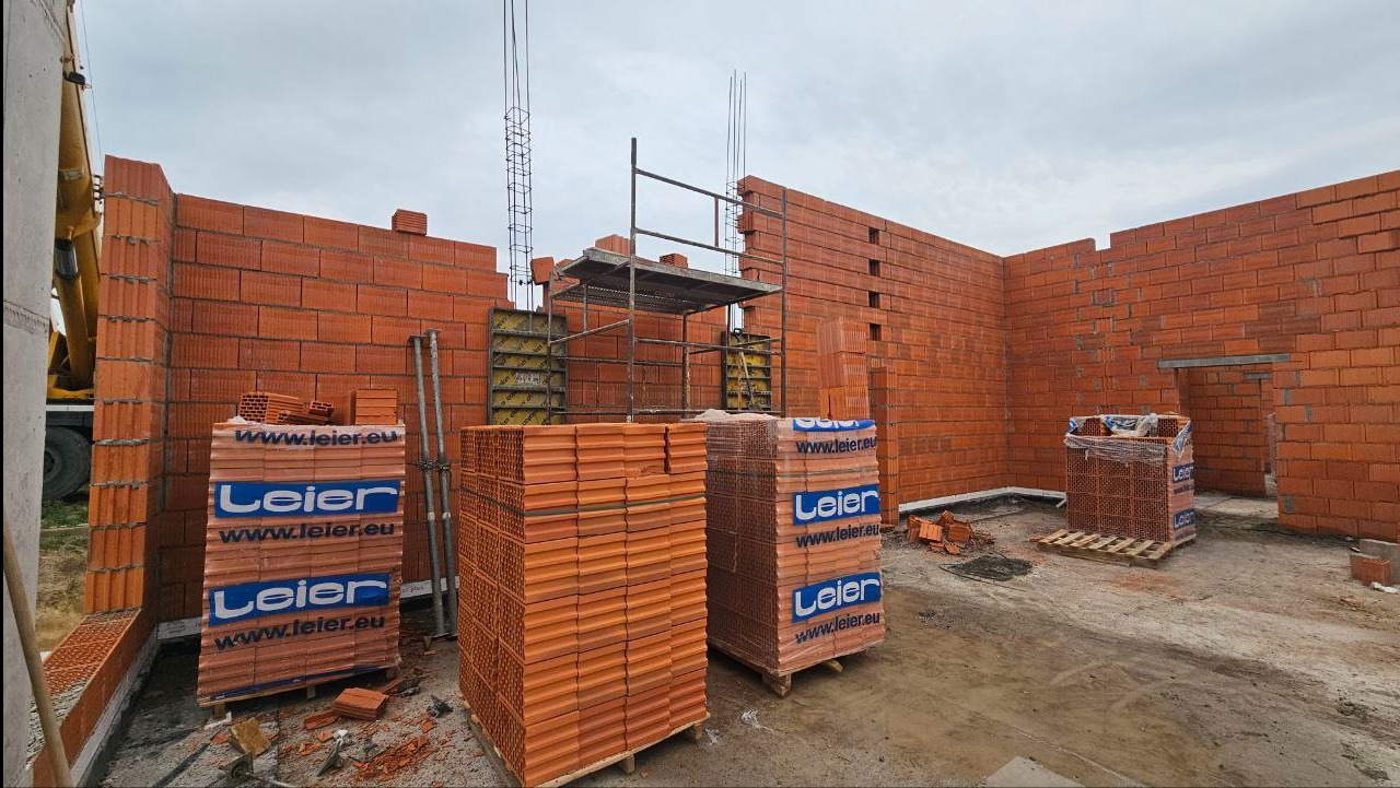 Wznoszenie ścian z czerwonej cegły Leier na placu budowy. Widoczne rusztowania, zbrojenie, palety z cegłami i fragment żurawia. Prace murarskie w toku.