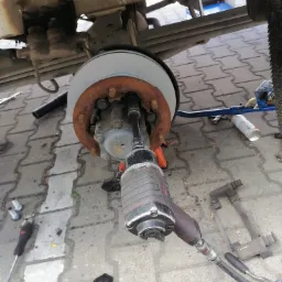 Wymiana tarczy hamulcowej w samochodzie ciężarowym, widoczny pneumatyczny klucz udarowy, nowa tarcza hamulcowa i skorodowane elementy mocujące.